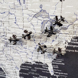 May include: A map of the United States with silver airplane push pins placed on various locations. The pins are all facing the same direction.