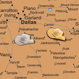 May include: A detailed map of Texas with two cowboy hat-shaped pins marking locations. The map is a warm brown color, with cities and towns labeled in black. The pins are metallic and add a decorative touch.