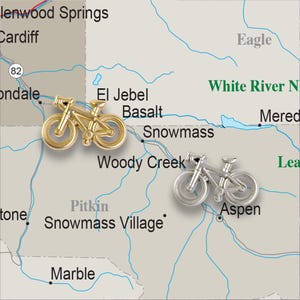 May include: A map of a region in Colorado with the names of towns and cities, including Aspen, Snowmass, and Basalt. Two small bicycle figurines are placed on the map, one gold and one silver.
