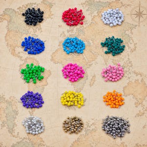 May include: A collection of fifteen distinct piles of round-headed push pins arranged in a grid. The colors include black, red, white, blue, light blue, dark green, bright green, hot pink, light pink, purple, yellow, orange, pearl, gold, and silver.