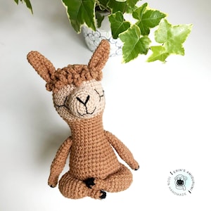 Op de afbeelding: Een bruine gehaakte lama die in meditatieve houding zit met zijn ogen dicht. De lama heeft een pluche bruine manen en een beige gezicht met zwarte ogen en een glimlach.