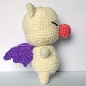 May include: A crocheted plush toy with a white body, purple wings, and a pink nose. The toy is standing on a white surface.