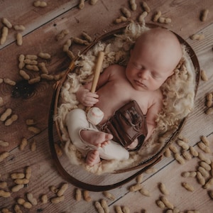 May include: A newborn baby lies in a woven basket on a wooden surface, surrounded by peanuts. The baby is nestled on a bed of fluffy, cream-colored material, holding a small baseball bat, a baseball, and a brown leather baseball glove.