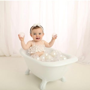 Puede incluir: Un bebé en una bañera blanca con patas de león llena de bolas blancas. El bebé lleva una diadema blanca y una parte superior blanca. Sostiene dos bolas transparentes. El fondo es blanco.