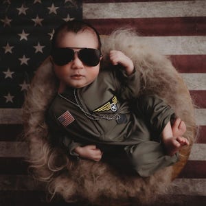 May include: A baby in aviator sunglasses and a green jumpsuit with a US flag patch and a wing emblem, on a fluffy surface against an American flag backdrop.