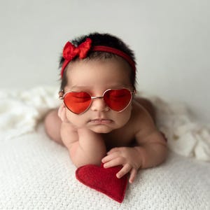 May include: A baby wearing red heart-shaped sunglasses and a red bow headband is lying on a white blanket. The baby is holding a red heart-shaped pillow.