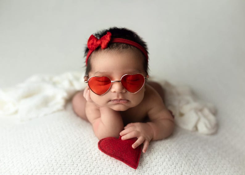May include: A baby wearing red heart-shaped sunglasses and a red bow headband is lying on a white blanket. The baby is holding a red heart-shaped pillow.