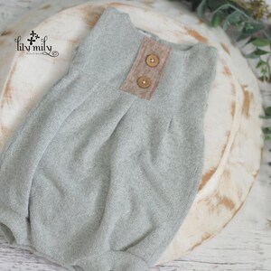 May include: A light gray baby romper with two brown buttons and a tan patch detail. The romper is made of a soft, textured fabric and is displayed on a wooden bowl. The background includes greenery.