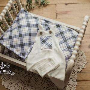 May include: A white waffle-knit baby romper with a plaid pocket and wooden buttons rests in a white wooden cradle. A plaid pillow in blue, white, and tan sits beneath the romper. The cradle is on a wooden surface.