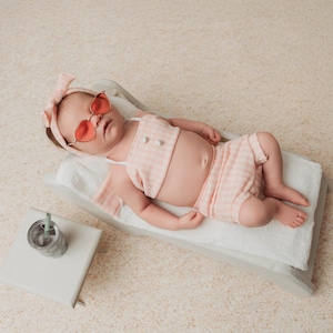May include: A baby wearing a pink gingham two-piece outfit, sunglasses, and a matching headband, lounging on a miniature beach chair. A small drink with a straw sits nearby. The scene is set on a neutral, textured surface.