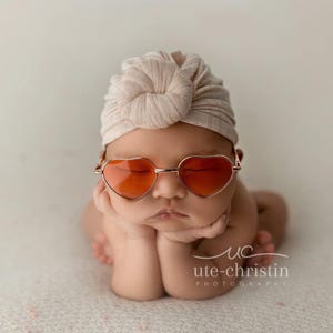 May include: A baby wearing a light brown turban and heart-shaped sunglasses with orange lenses. The baby is sitting with their hands clasped in front of them.