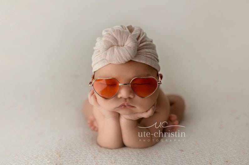 May include: A baby wearing a light brown turban and heart-shaped sunglasses with orange lenses. The baby is sitting with their hands clasped in front of them.