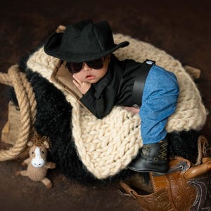 May include: A newborn baby dressed in a black cowboy hat, black sunglasses, a black long-sleeved shirt, blue jeans, and black boots. The baby rests on a cream-colored chunky knit blanket with a small brown knitted horse toy and a brown leather saddle.