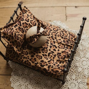 May include: A miniature black metal bed with a leopard print pillow and blanket. A round, cream-colored prop with a matching leopard print bow is on the bed. The bed is on a cream-colored lace doily, set on a wooden surface.
