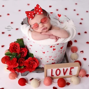 May include: A newborn baby wearing red and white polka dot sunglasses and a red and white polka dot bow is sleeping in a white bucket with a pink and white heart pattern. The bucket is sitting on a white surface with red hearts scattered around it. There is a bouquet of red roses and a wooden sign that says "LOVE" in red letters.