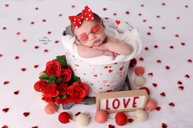 May include: A newborn baby wearing red and white polka dot sunglasses and a red and white polka dot bow is sleeping in a white bucket with a pink and white heart pattern. The bucket is sitting on a white surface with red hearts scattered around it. There is a bouquet of red roses and a wooden sign that says "LOVE" in red letters.