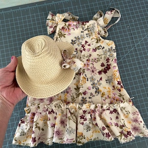 May include: A floral print dress and matching straw hat. The dress features a cream background with purple, yellow, and gray flowers. The hat is a light tan color with a matching floral accent. The dress has a ruffled hem and shoulder ties.