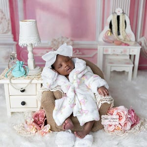 May include: A sleeping baby wearing a white robe with pink floral accents and a large white bow. The baby is seated in a small brown chair, surrounded by decorative elements including a white lamp, a small table, and pink roses.