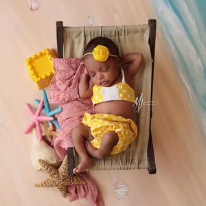 May include: A baby wearing a yellow and white polka dot swimsuit with a yellow flower headband is sleeping on a small bed with a pink blanket and starfish decorations.
