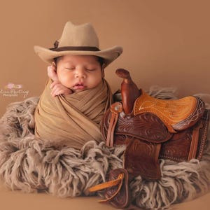 May include: A newborn infant wrapped in tan fabric, wearing a beige cowboy hat, resting in a fluffy gray nest next to a miniature brown leather saddle with intricate floral tooling and a light tan seat, all set against a solid tan background.