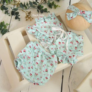 Puede incluir: Un conjunto de ropa de bebé con estampado floral azul claro, que incluye una blusa con volantes, un bloomer y una diadema a juego con un lazo. El conjunto se exhibe sobre un soporte de madera blanco con vegetación de fondo. El texto "Back Side" es visible.