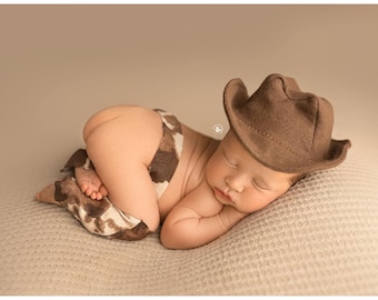 May include: A brown felt cowboy hat and a pair of brown and white cow print chaps worn by a sleeping infant. The hat features a classic curved brim and a pinched crown, while the chaps are designed with a spotted pattern covering the legs.