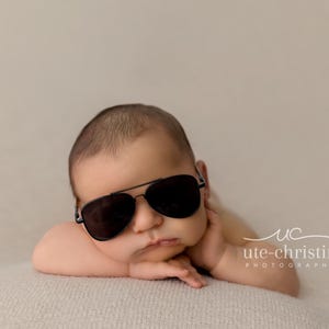 May include: A baby wearing black sunglasses is lying on a light brown blanket. The baby is looking at the camera with a serious expression.