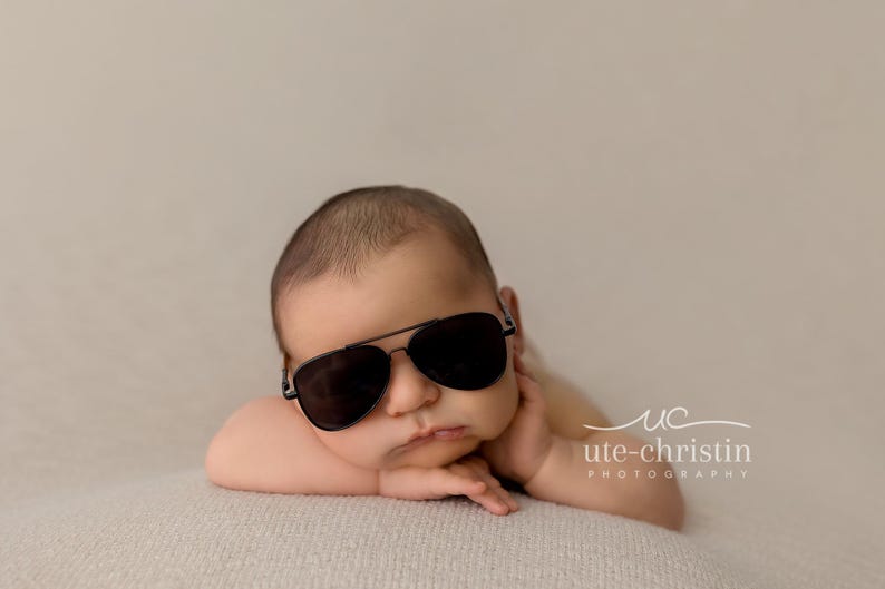 May include: A baby wearing black sunglasses is lying on a light brown blanket. The baby is looking at the camera with a serious expression.