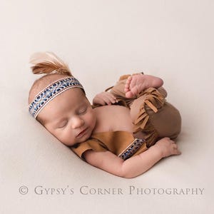 May include: A newborn baby wearing a Native American-inspired outfit, including a fringed pants, a top, and a headband with a feather. The baby is lying on a soft, cream-colored surface.