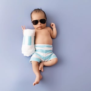 May include: A newborn infant wearing black-framed sunglasses and light blue and white striped shorts, holding a rolled-up white towel with light blue stripes. The infant is positioned against a solid, light purple background.