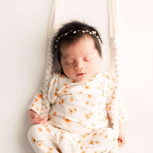 May include: A sleeping baby wearing a floral print outfit and a pearl headband, resting on a small, white swing. The outfit is cream-colored with orange flower patterns. The swing is suspended by cream-colored macrame ropes.