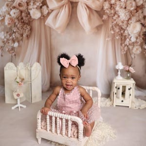 May include: A baby girl in a pink dress sits on a small white bed. She wears a pink bow headband. The background features a large bow, floral arrangements, and a small side table with a lamp.