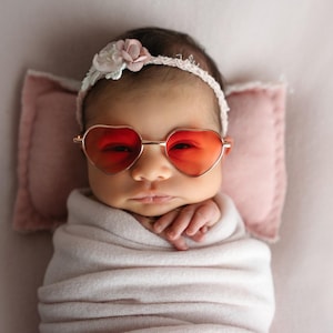 May include: A baby wearing a white headband with pink flowers and heart-shaped sunglasses. The baby is wrapped in a white blanket and lying on a pink surface.