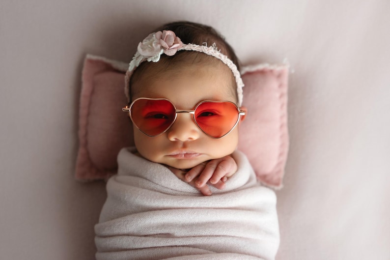 May include: A baby wearing a white headband with pink flowers and heart-shaped sunglasses. The baby is wrapped in a white blanket and lying on a pink surface.
