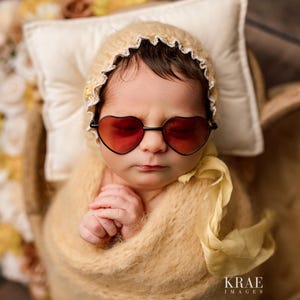 May include: A baby wearing a white crocheted hat with a yellow crocheted blanket and heart-shaped sunglasses.