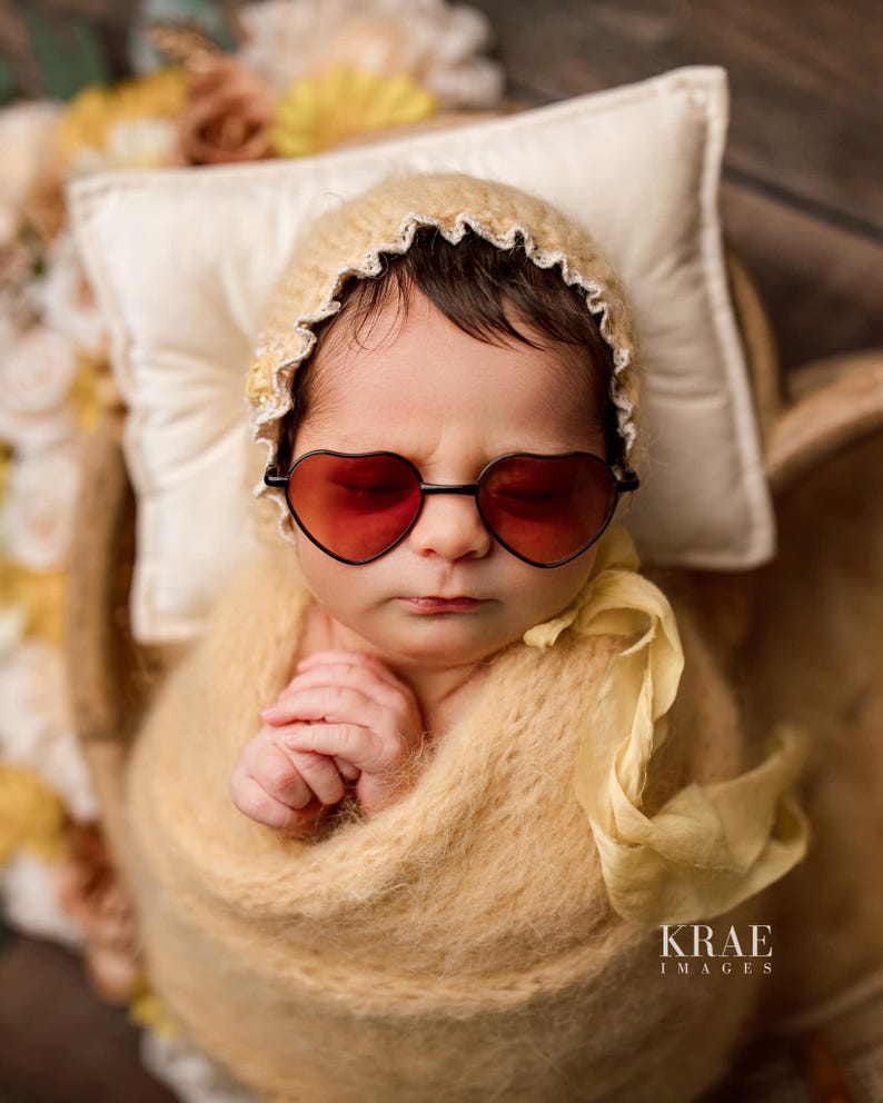 May include: A baby wearing a white crocheted hat with a yellow crocheted blanket and heart-shaped sunglasses.