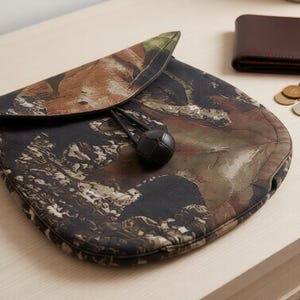May include: A camouflage pattern pouch with a drawstring closure, alongside a brown leather wallet, keys, and coins. The pouch features a mix of brown, green, and black hues. A black cat illustration is in the lower left corner.