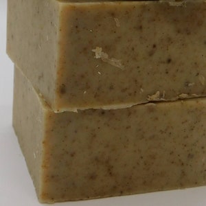 May include: Two stacked bars of handmade soap. The soap has a light brown color with darker brown speckles throughout. The edges are slightly rough, and the soap appears to be unscented. The bars are rectangular and have a natural, rustic appearance.