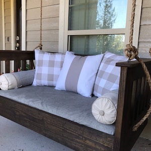 May include: A dark brown wooden porch swing with a gray cushion and white and gray patterned pillows. The swing is hanging from a porch ceiling with thick rope.