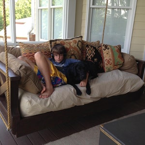 May include: A wooden swing bed with a beige mattress and colorful patterned pillows. A young person is sitting on the bed with a black dog. The swing bed is hanging from the ceiling of a porch.