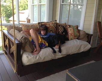 May include: A wooden swing bed with a beige mattress and colorful patterned pillows. A young person is sitting on the bed with a black dog. The swing bed is hanging from the ceiling of a porch.