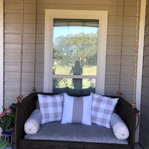 May include: A wooden swing bed with a gray cushion and pillows. The pillows have a white and gray checkered pattern and a white and gray striped pattern. The swing bed is hanging from the ceiling of a porch.