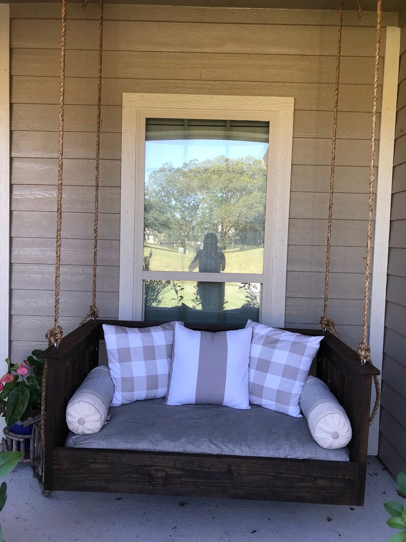 May include: A wooden swing bed with a gray cushion and pillows. The pillows have a white and gray checkered pattern and a white and gray striped pattern. The swing bed is hanging from the ceiling of a porch.