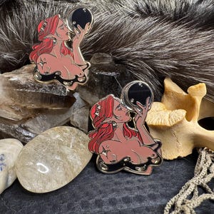 Könnte beinhalten: Zwei Emaille-Pins, die eine Frau mit roten Haaren zeigen, die eine schwarze Kugel hält. Die Frau ist mit nackter Brust und einem fließenden roten Gewand dargestellt. Die Pins sind vor einem Hintergrund aus Kristallen und Fell platziert.