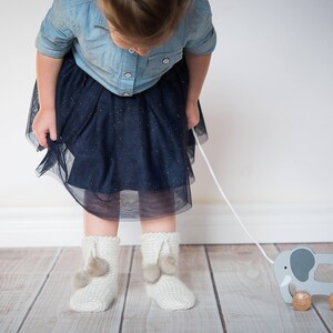 Pode incluir: Uma menina pequena vestindo uma camisa de brim azul e uma saia de tule preta puxa um elefante de brinquedo de madeira cinza sobre rodas com uma corda branca.