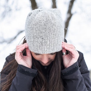 Könnte beinhalten: Eine Frau trägt eine graue Strickmütze und eine schwarze Jacke mit Kapuze. Sie steht in einer schneebedeckten Umgebung.