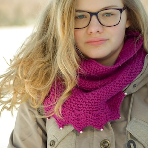 May include: A young woman with long blonde hair wearing a pink crocheted scarf and a brown jacket. She is wearing glasses with black frames.