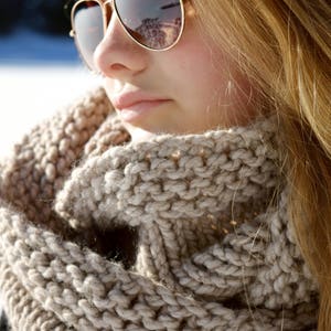 May include: A close-up of a woman wearing a beige knitted scarf and sunglasses. The scarf is thick and warm, and the sunglasses are stylish and modern.