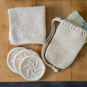 Puede incluir: Un conjunto de artículos tejidos a mano, de color crema, sobre una superficie de madera. El conjunto incluye un paño de lavado, una bolsa para jabón, discos faciales reutilizables y una pastilla de jabón con remolinos azules.