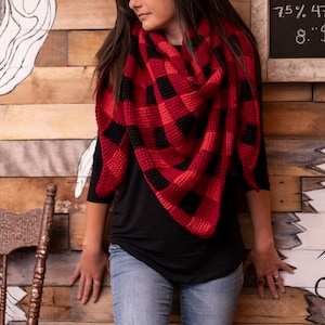 May include: A red and black plaid scarf with a fringe edge. The scarf is worn by a person standing in front of a wooden wall with a white drawing on it.